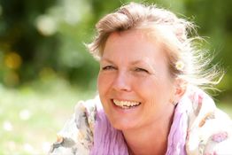 Ältere Frau lacht im Freien bei sonnigem Wetter, umgeben von unscharfem Grün im Hintergrund.