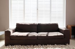 Braunes Sofa vor Fenster mit Jalousien, Sonnenlicht fällt durch Lamellen auf den Teppich.