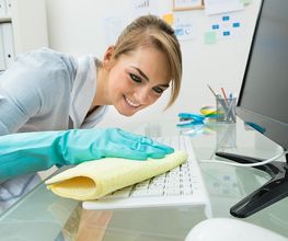 Frau reinigt Tastatur am Schreibtisch mit gelbem Tuch, trägt blaue Handschuhe und lächelt.