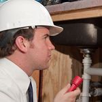 Person in a hard hat examining pipes with a flashlight under a sink.