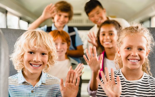 Kinder sitzen lachend in einem Reisebus auf Klassenfahrt. Sie winken in die Kamera.