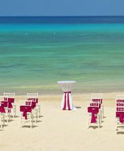 Sillas blancas en la playa, listones rojos y mesa alta, con vista al mar azul.