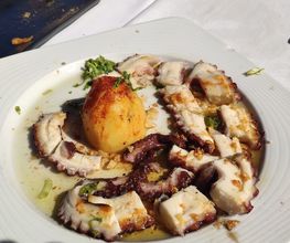Plato de pulpo a la plancha con una papa asada en un plato blanco sobre mesa al aire libre.