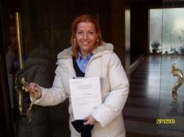 Donna sorridente apre una porta, tenendo dei documenti in mano, indossa un cappotto bianco.