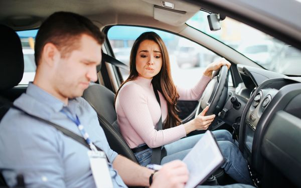 Eine Fahrschülerin mit Prüfungsangst sitzt neben ihrem Prüfer im Auto. Der Prüfer hält ein Klemmbrett mit Bewertungsbogen in der Hand, den er konzentriert ausfüllt.