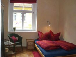 Kleines Schlafzimmer mit Fenster, rotes Bettzeug, Stuhl und Tisch mit Pflanze und Zeitschriften.