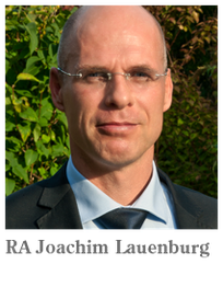 Ein Mann mit Anzug und Brille steht vor einem grünen Hintergrund im Freien.