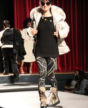 Person auf Laufsteg mit Sonnenbrille, Felljacke, gemusterten Leggings vor rotem Vorhang.