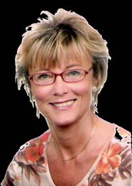 Eine lächelnde Frau mit Brille und Blumenbluse vor schwarzem Hintergrund.