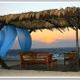 Holzlaube am Strand mit blauen Vorhängen, Sonnenuntergang im Hintergrund.