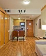 Sala de estar com sofá, mesa de jantar e cozinha compacta ao fundo, em tons de madeira clara.