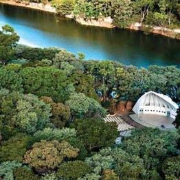 Vista aérea de parque verde com um lago e uma estrutura branca de telhado curvo entre as árvores.