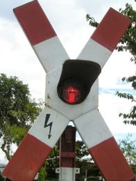Rotes Licht an einem Eisenbahnkreuzungssignal mit weiß-rotem Andreaskreuz bei Tageslicht.