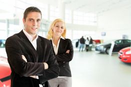 Menschen in Businesskleidung stehen vor Autos in einem hellen, modernen Autohaus.