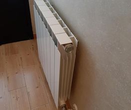Radiador blanco en una pared de habitación con suelo de madera clara y cortina blanca al fondo.