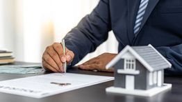 Person signing a document near a small house model on a desk, symbolizing real estate transaction.