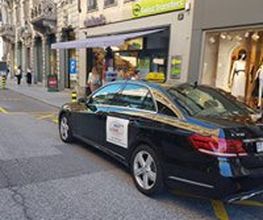 Schwarzes Auto parkt auf einer Stadtstraße vor Geschäften mit Passanten im Hintergrund.