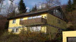Gelbes Haus mit braunem Dach und Balkon, umgeben von Bäumen, am Waldrand.