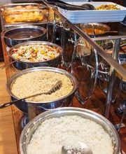 Buffet com pratos variados, incluindo arroz, farofa e salada, em tigelas de aço inoxidável.