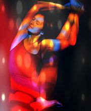 Person tanzt in buntem Licht, trägt ein rotes Outfit und hat die Arme über den Kopf ausgestreckt.