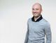 Ein lächelnder Mann in grauem Pullover vor weißem Hintergrund.