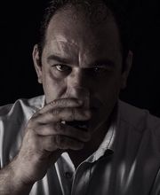 Mann in Hemd trinkt aus einem Glas und schaut ernst. Hintergrund ist dunkel.