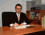 Ein Mann in Anzug sitzt am Schreibtisch, schreibt auf einem Papier und lächelt leicht.