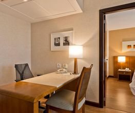 Escritório em hotel com mesa, cadeira e lâmpada; quarto visível ao fundo.