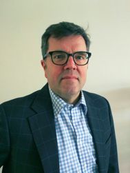 Man met bril en geruit overhemd in donkerblauw jasje voor een witte achtergrond.