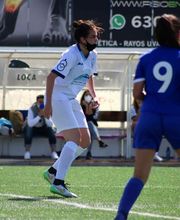 Jugadoras de fútbol en partido, una de blanco y otras de azul, en un campo con banquillo detrás.