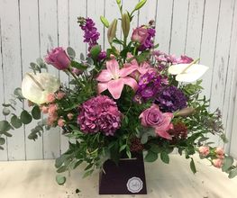 Ramo de flores variadas en tonos rosados y lilas sobre fondo de madera blanca.