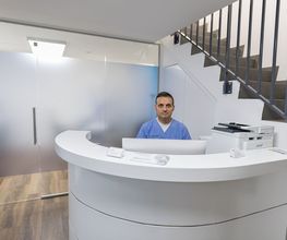 Mann in blauer Kleidung an weißem Empfangstresen, Treppe und Glaswand im Hintergrund.