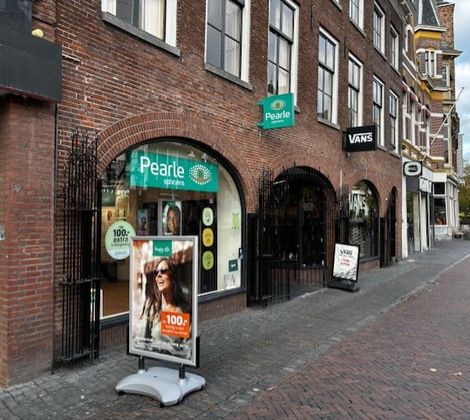Winkelstraat met Pearle opticien en andere winkels, voetgangers op trottoir, bewolkte lucht.