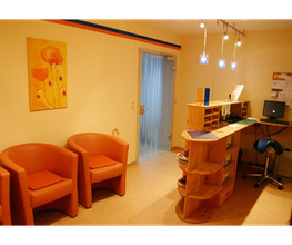 Wartezimmer mit drei orangefarbenen Sesseln, Holztresen, gelber Wand und Mohnblumenbild.