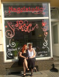 Zwei Frauen sitzen vor einem dekorierten Nagelstudio. Sie lachen und tragen Sommerkleidung.