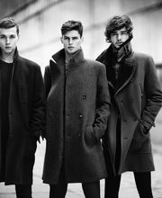 Tre uomini in cappotti eleganti posano all'aperto in bianco e nero.