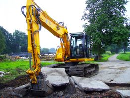 Ein gelber Bagger reißt einen Asphaltweg auf, umgeben von grünen Bäumen und Wiesen.