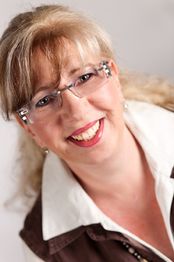 Eine lächelnde Frau mit Brille und blondem Haar vor einem neutralen Hintergrund.