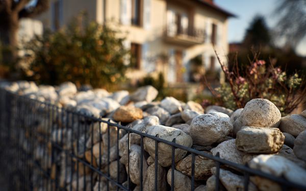 Gabione befüllt mit buntem Steinbruch. Im Hintergrund ist ein beiges Einfamilienhaus mit Fensterläden zu sehen.