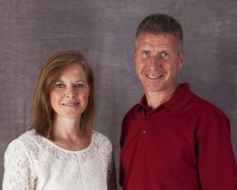 Man in red shirt and woman in white lace top standing against a gray backdrop.