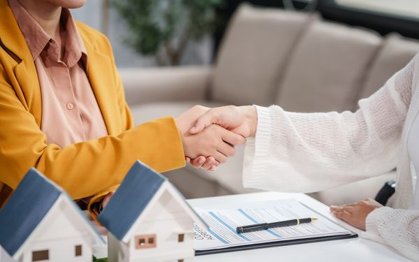 Zwei Personen schütteln sich die Hände. Auf dem Tisch vor ihnen liegt ein Klemmbrett mit Kugelschreiber. Symbolisch stehen im Vordergrund zwei Modellhäuser.