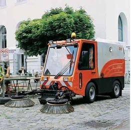 Orange Straßenkehrmaschine auf gepflastertem Platz vor einem Gebäude mit Bäumen im Hintergrund.