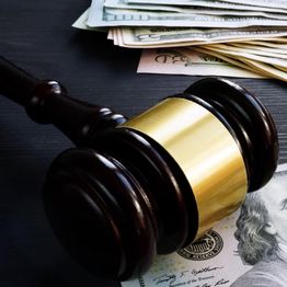 Gavel on a hundred-dollar bill, with a stack of various banknotes in the background.