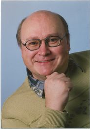 Eine lächelnde Person mit Brille und grünem Pullover, die den Kopf auf eine Hand stützt.