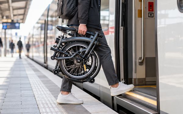Ein Pendler trägt ein zusammengeklapptes Klapprad am Bahnhof und steigt damit in einen Zug ein, um das kompakte Faltrad auf dem Arbeitsweg als Handgepäck mitzunehmen.