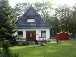 Haus mit Giebeldach, Garten, Liegestuhl und rotem Schuppen im Hintergrund, umgeben von Bäumen.