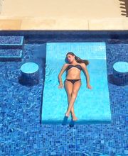Mujer en bikini descansa sobre flotador azul en piscina con mosaicos y columnas alrededor.