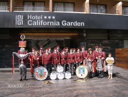 Blaskapelle in roten Uniformen vor dem Hotel California Garden, Trommeln und Instrumente dabei.