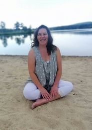 Frau sitzt lächelnd im Schneidersitz am Strand vor einem See mit Bäumen im Hintergrund.
