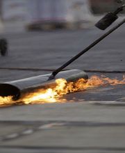 Brenner erhitzt Bitumenrolle auf Dachoberfläche zur Abdichtung.